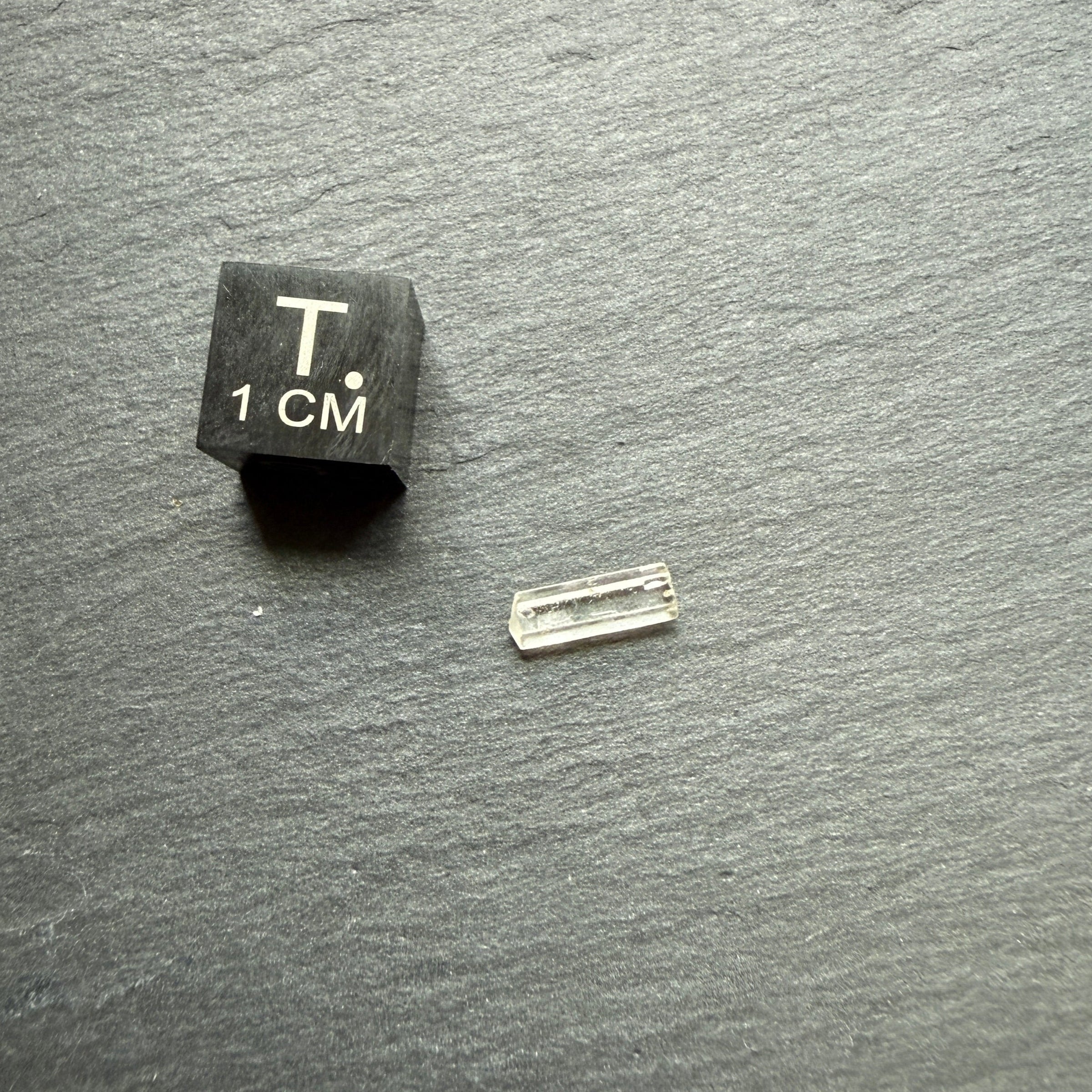 Small black cube with 'T' and '1 cm' marking next to a clear crystal on a dark surface